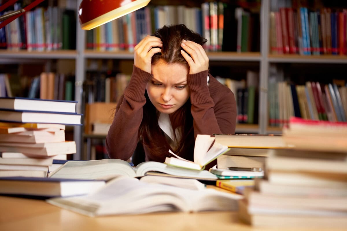 A young woman sits at a desk in a library, holding her head in frustration while surrounded by open books and stacks of papers, indicating stress or difficulty studying.