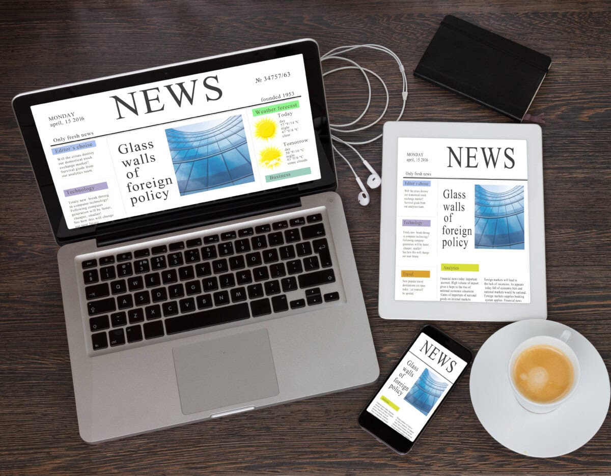 A laptop, tablet, and smartphone on a wooden desk display the same news website. Also visible are headphones, a notebook, a pen, and a cup of coffee on a saucer.
