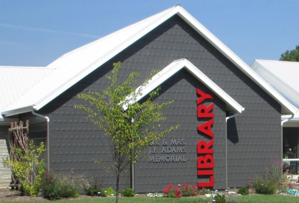 A modern gray building with a sloped white roof has large red vertical letters spelling LIBRARY on the side. Smaller letters read J.E. & MRS. J.F. ADAMS MEMORIAL. A young tree and flowerbed are in front.