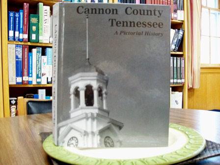 A book titled Cannon County Tennessee: A Pictorial History stands upright on a green decorative plate in a library, with bookshelves and a window in the background.