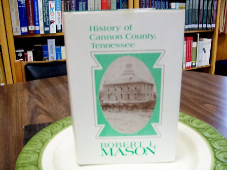 A book titled History of Cannon County, Tennessee by Robert L. Mason stands upright on a plate, with bookshelves full of books in the background. The cover features a green border and an old building photo.