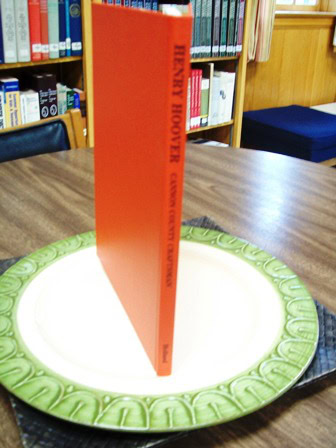 An orange book titled Henry Hoover stands upright on a decorative plate with a green border, placed on a wooden table in a library setting.