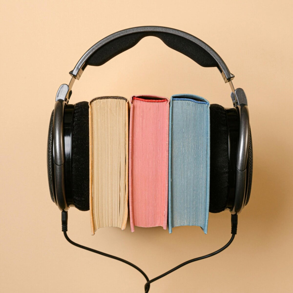 A pair of black headphones is placed over three upright books—one beige, one pink, and one blue—against a light tan background. The setup suggests the concept of audiobooks.